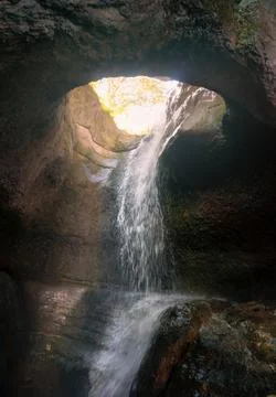 Waterfall inside a dark cave flowing through a stone hole with sunlight Stock Photos
