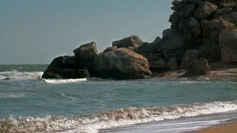 The wave breaks on the sandy beach, with a mountain of boulders and rocks in the Stock Footage 220998468