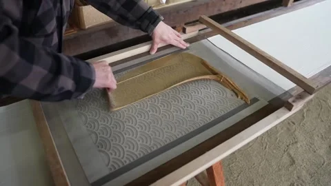 Wave patterned stencil screen lowered onto white cloth as the dyer steadies the Stock Footage 342319476