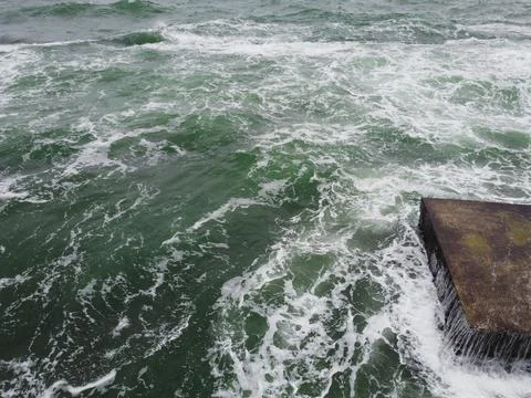 The wave rolls over a large square rock lying on the shore, covered with green Foto stock