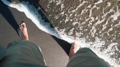 Wave washes over man's legs in shorts on black sand Stock Footage 63529127