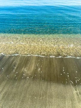 Waves break on sandy shore under clear sky with calm water reflecting sunli.. Stock Photos