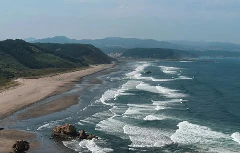 Waves breaking on a long, sandy beach. The coastline is defined by green, rol Stock Photos
