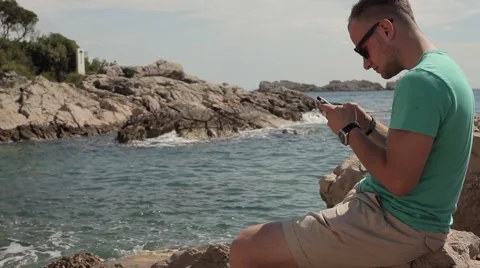 Waves breaking the rocks. Young man with mobile phone Stock Footage 55281536