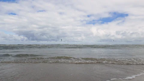 Waves breaking on sandy beach with cloudy sky and distant kite surfers Stock Footage 346641751