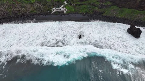 Waves continuously crash onto a black sand beach, revealing rocky cliffs Stock Footage 328523549