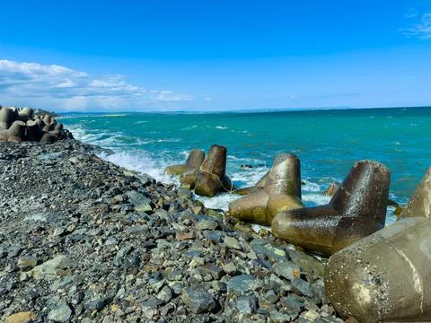 Waves crash against breakwater structures near rocky shore under blue sky a.. Stock Photos