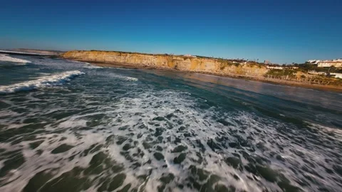 Waves crash against golden cliffs under clear blue skies Stock Footage 339787226