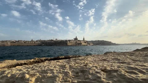 Waves crash against golden limestone rocks valletta skyline rises across water Видео 343677401