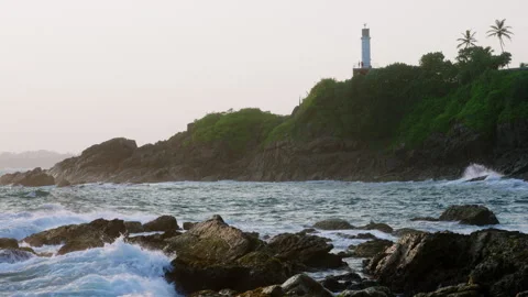Waves crash against rocks at sunset with lighthouse on a hill. Video captures Stock Footage 272173590
