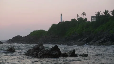 Waves crash against rocks at sunset with lighthouse on hill. Twilight ocean Stock Footage 273747550