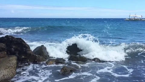 The waves crash in on the black rocks in the water. A boat is floating in Vídeos de archivo 264906589