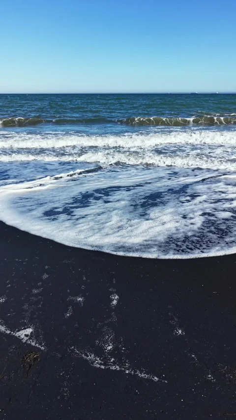 Waves crash on a black sand beach with smooth stones on a sunny day near the Stock Footage 335389160