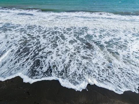 Waves crash on a black sand beach under a clear sky, showing the interactio.. Stock Photos