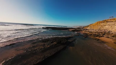 Waves crash on dark, rocky shore under clear blue sky Stock Footage 339787066