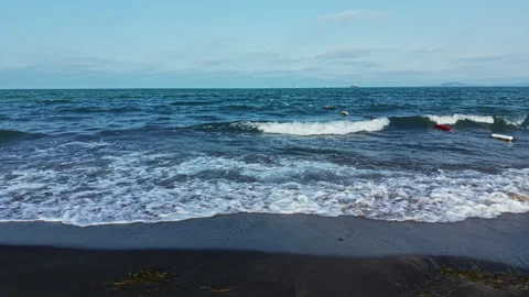 Waves crash on a dark sandy beach with distant ships and a clear blue sky in the Stock Footage 337883915