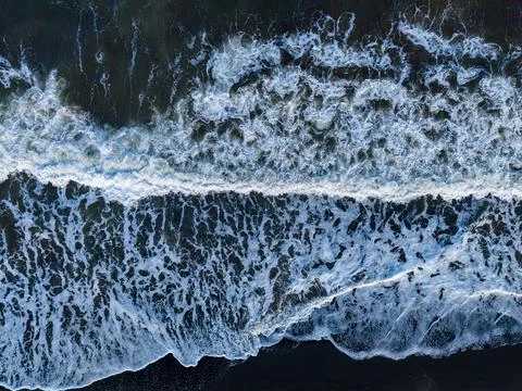 Waves crash on a dark sandy beach during daylight, creating a pattern of wh.. Stock Photos