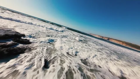 Waves crash over dark rocks under bright blue sky Stock Footage 339786234