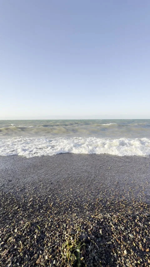 Waves crash on a pebble beach under a clear blue sky Stock Footage 338347718