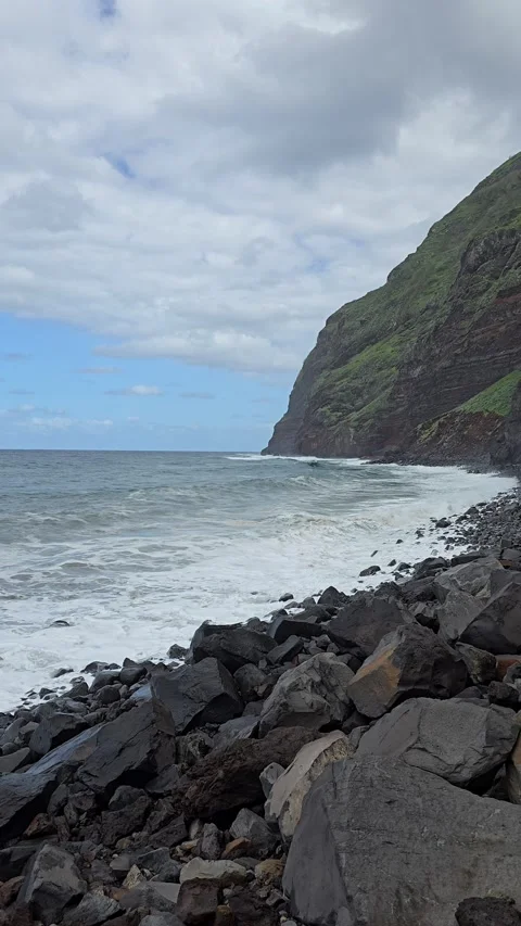 Waves crash on the rocky shore beneath towering green cliffs of Madeira’s  Stock Footage 310024416