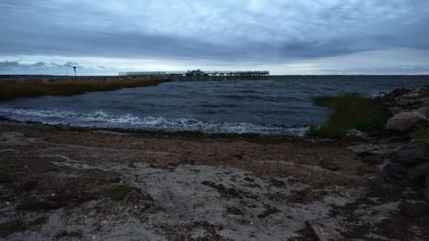 Waves crash on a rocky shore with a distant pier under a cloudy sky Vídeo Stock 346097902