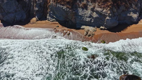 Waves crash on a sandy beach next to rocky cliffs during a sunny day Stock Footage 344811145