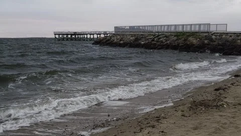 Waves crash on sandy beach with pier and modern structure in background Vídeo Stock 346097843