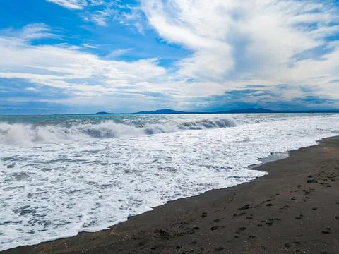 Waves crash on sandy beach under blue sky with clouds and distant mountains.. Stock Photos