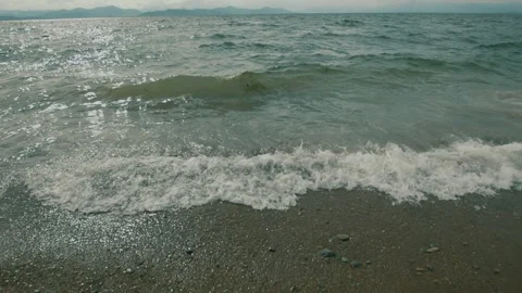 Waves crash onto shore creating foam at edges and washing pebbles out of sand Видео 318671332