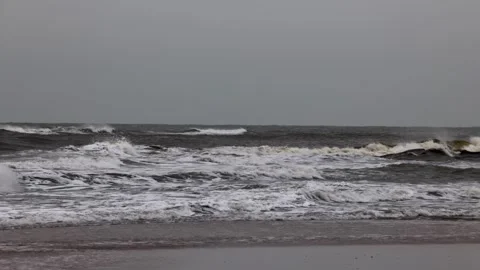 Waves crash onto the shore under a gray, overcast sky. The sea appears rough Stockbeeldmateriaal 305202645