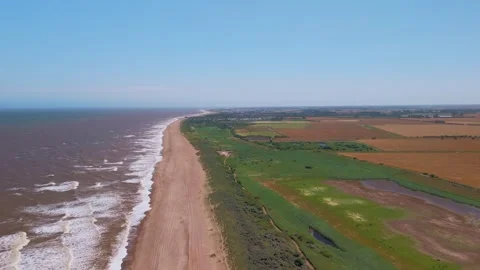 Waves crash onto wide sandy beach under clear blue sky. Fields stretch beyond Stock Footage 342025498