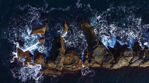 Waves crashing against rugged rocks on a serene coastline at sunset in an Stock Footage 331984574