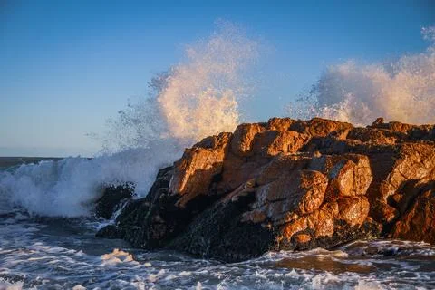 Waves crashing dramatically against rugged coastal rocks at sunset. Stock Photos