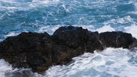 Waves crashing over rugged black volcanic rocks, creating white foam, splashes Видео 346247244