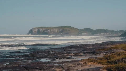 Waves crashing on a rocky beach with a distant cliff and town under a clear sky Stock Footage 341321787