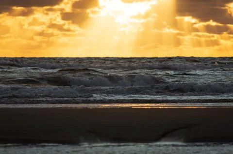 Waves crashing on sand dune while the sun breaks to the clouds Stock Photos