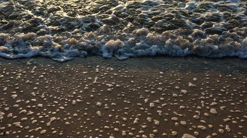 Waves crashing on the shore create foam and bubbles at sunset by the beach Stock Photos
