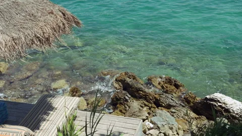 Waves crushing of beachfront wooden deck with sun loungers above rocky turq.. Stock Footage 338869672