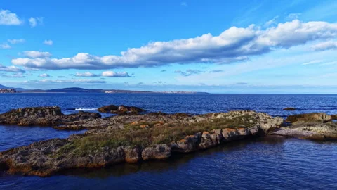 Waves gently lap against rocky shores under a clear blue sky with fluffy clouds Stock Footage 336590693