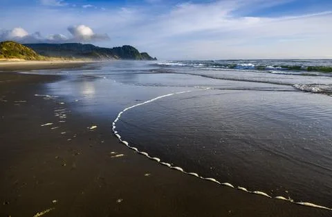 Waves gently lap the sandy shore at sunset near a tranquil coastal landscape Stock Photos