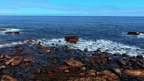 Waves hitting large red rock surrounded by coastal boulders. Sea swirling over Stock Footage 314267903