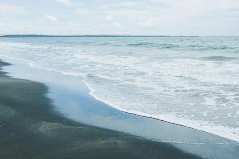 Waves move gently onto the sand at a beach with a clear sky and distant land  Foto stock