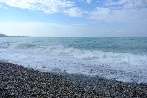 Waves on a pebble beach. White clouds in the blue sky over the sea. Stock Photos