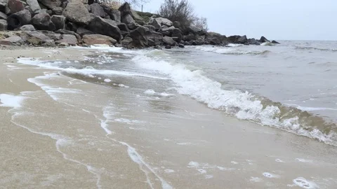 Waves roll ashore leaving foam behind. The gray landscape and horizon create a s Stock-Footage 120627576