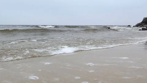 Waves roll ashore leaving foam behind. The gray landscape and horizon create a s Stock Footage 120627664