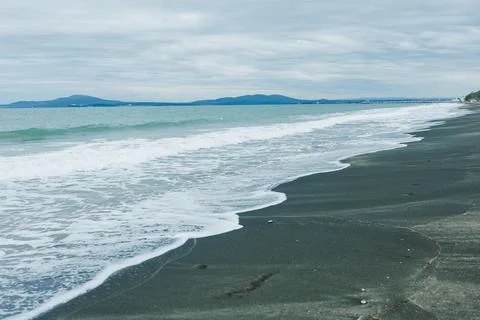 Waves roll onto a black sand beach while distant islands are visible under a  Fotos Stock
