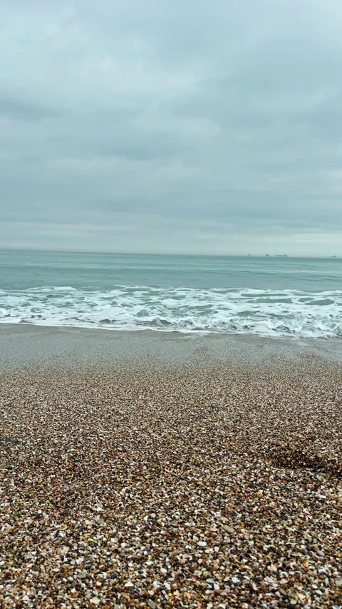 Waves roll onto a pebbled beach on an overcast day with distant ships visible on Stock Footage 340878796
