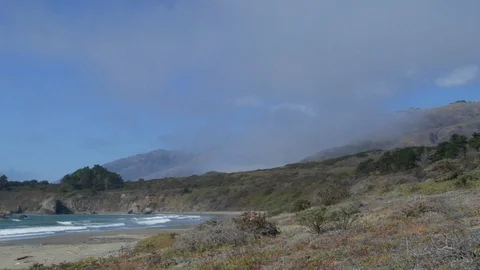 Waves roll into shore onto sand on empty beach with cliff and mountains fog Stock Footage 125341386