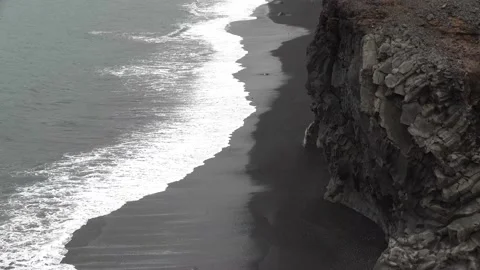 Waves rolling in over black sandy beach below steep cliffs on a cloudy day 1 Stock Footage 136616341