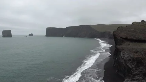 Waves rolling in over black sandy beach below steep cliffs and a stony arch Stock Footage 136616352
